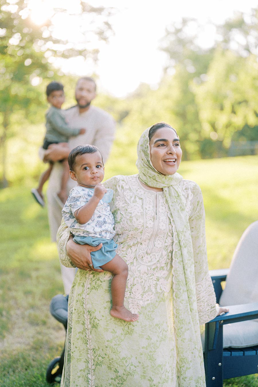 A smiling woman in a light green, embroidered dress holds a young child in a park, with another adult and child in the blurred background, surrounded by lush greenery and sunlight.