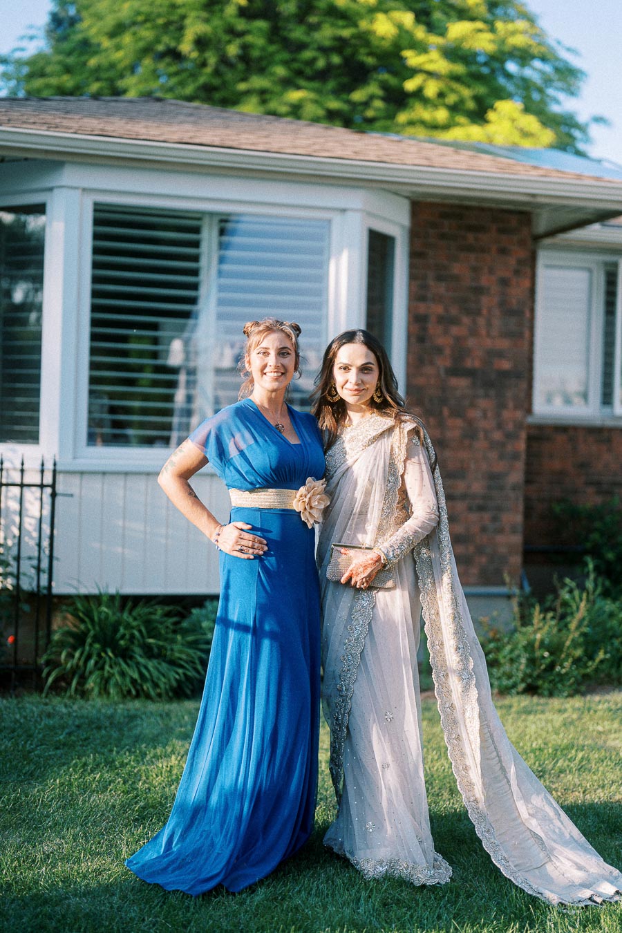 Two women smiling, one in a blue dress and the other in a cream traditional outfit, standing outdoors in front of a house with a garden background.