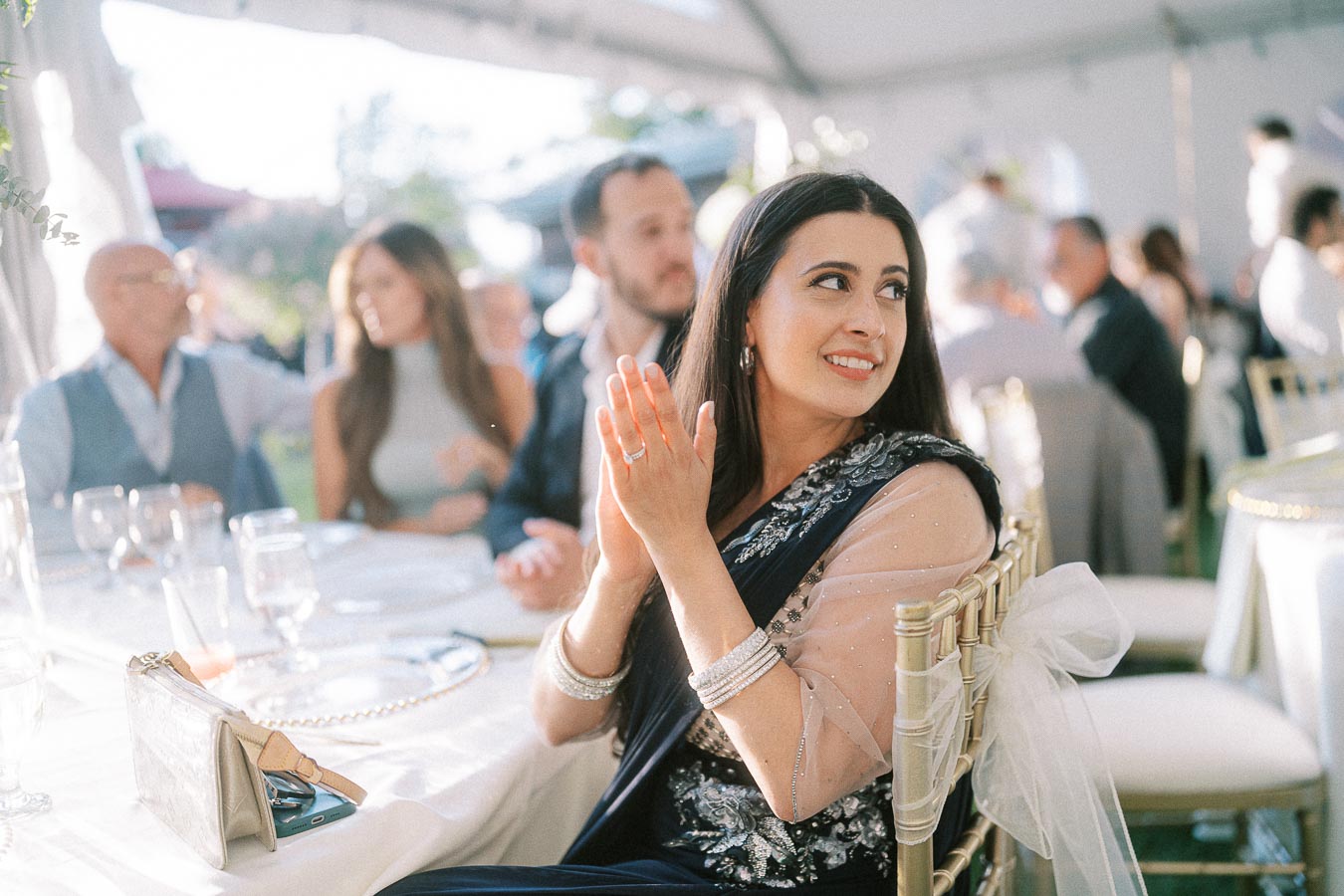 Woman clapping at a formal outdoor event, seated at a decorated table with elegant glassware and cloth napkins, wearing a dark, ornate outfit with shimmering details, surrounded by other attendees in a sunlit tent setting.