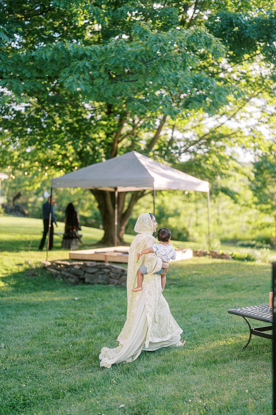 A woman in a light floral dress carrying a child outdoors in a sunny, green garden setting, with a canopy tent and tree in the background.
