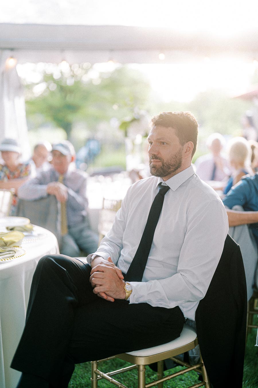 A man in formal attire with a white shirt and black tie sits attentively on a chair at an outdoor event, surrounded by other guests.