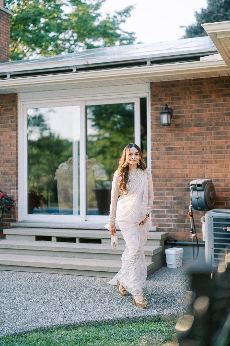 Woman in elegant traditional saree walking outdoors in a backyard setting with brick house in the background.