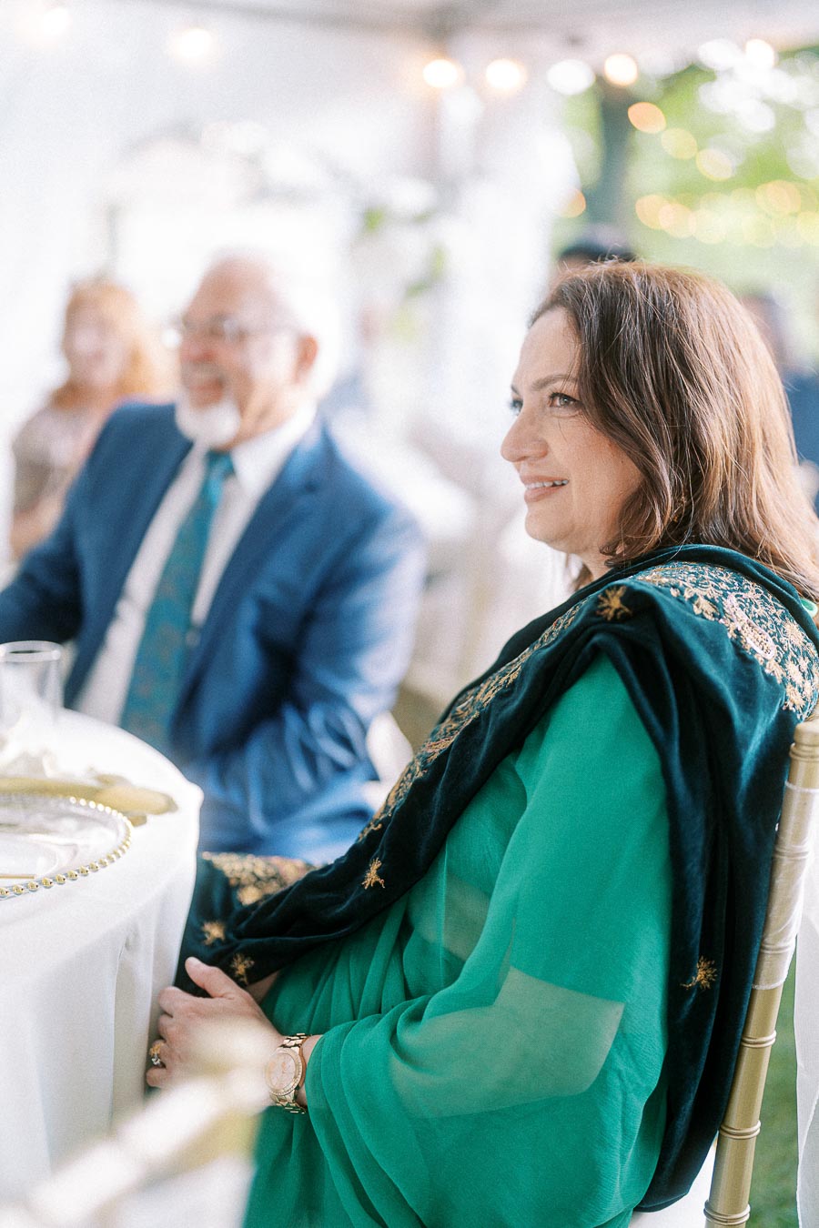 Elegant woman in green attire and black shawl smiling at a formal event, sitting at a table with blurred guests in the background.