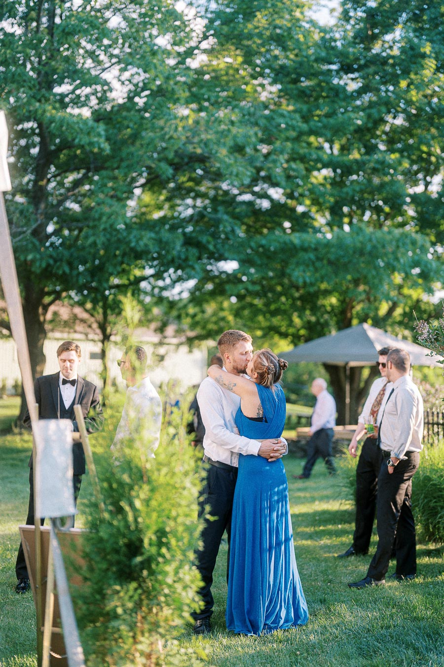 Couple embracing at an outdoor wedding reception, surrounded by guests in formal attire under a canopy of lush green trees.