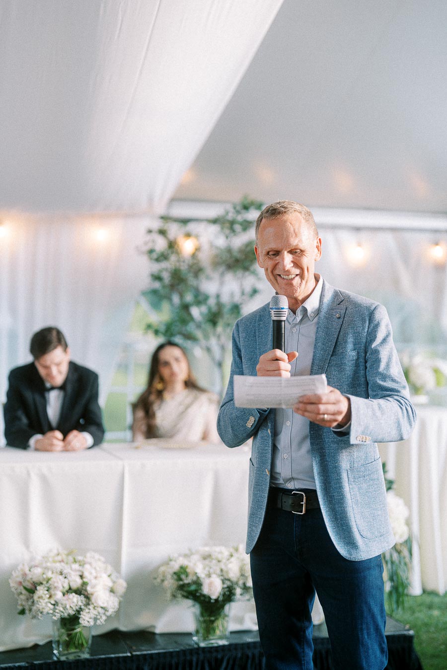 A man in a blue blazer giving a speech with a microphone at a formal indoor event, with two people seated at a table in the background.