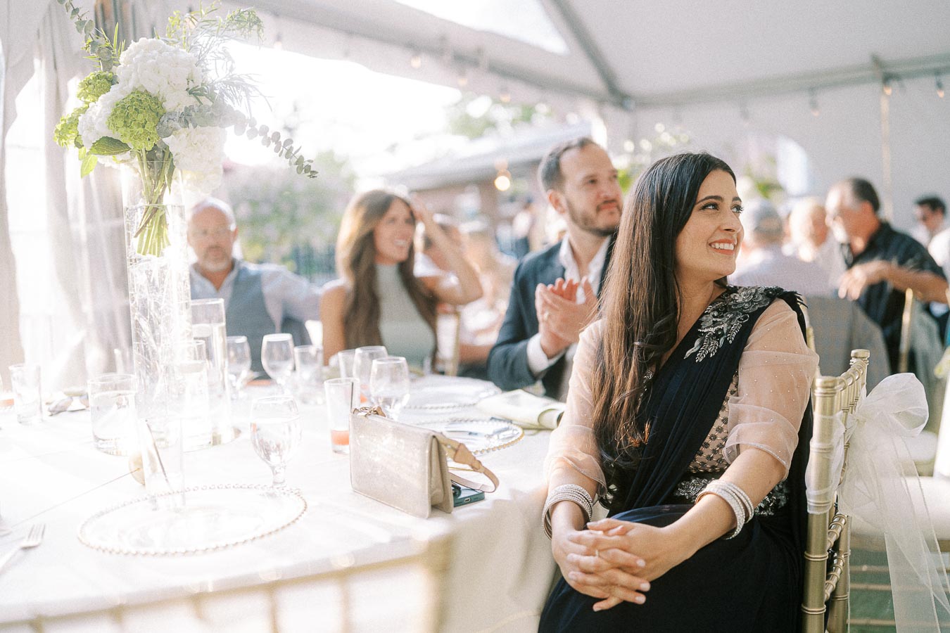 Group of elegantly dressed guests enjoying an outdoor wedding reception under a white canopy, featuring a smiling woman in a dark dress with floral embroidery, seated near a beautifully arranged table with flowers and glassware.