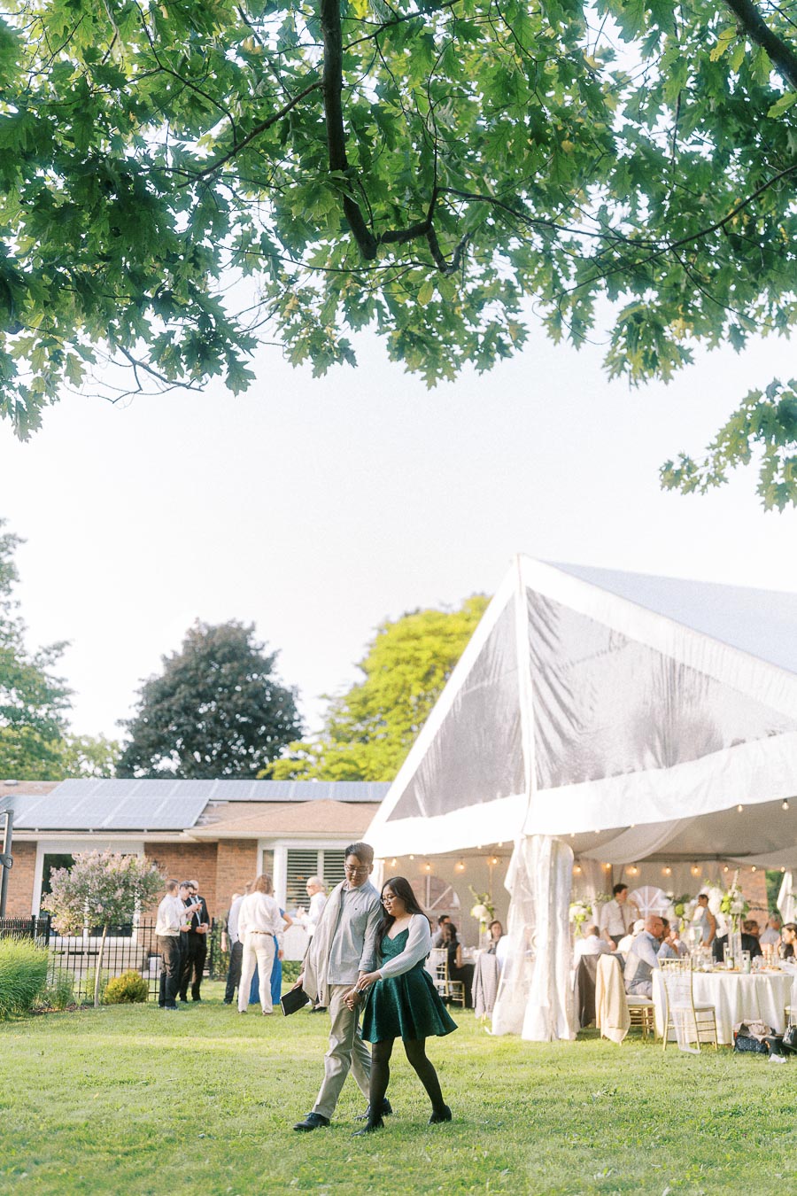 Outdoor wedding reception under a white tent with guests mingling on a sunny day, surrounded by lush greenery and a view of solar panels on a nearby building.