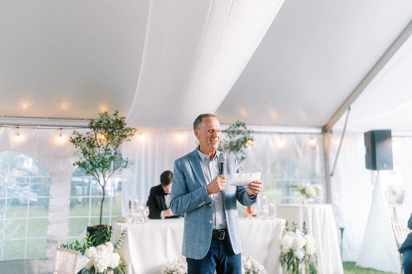 Man in a blue blazer giving a speech at a wedding reception in a bright, decorated tent with flowers and greenery.