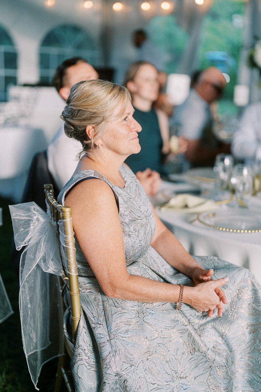 A woman in a formal dress sits attentively at a wedding reception, surrounded by elegantly set tables and soft lighting, capturing the joyful ambiance of a special event.