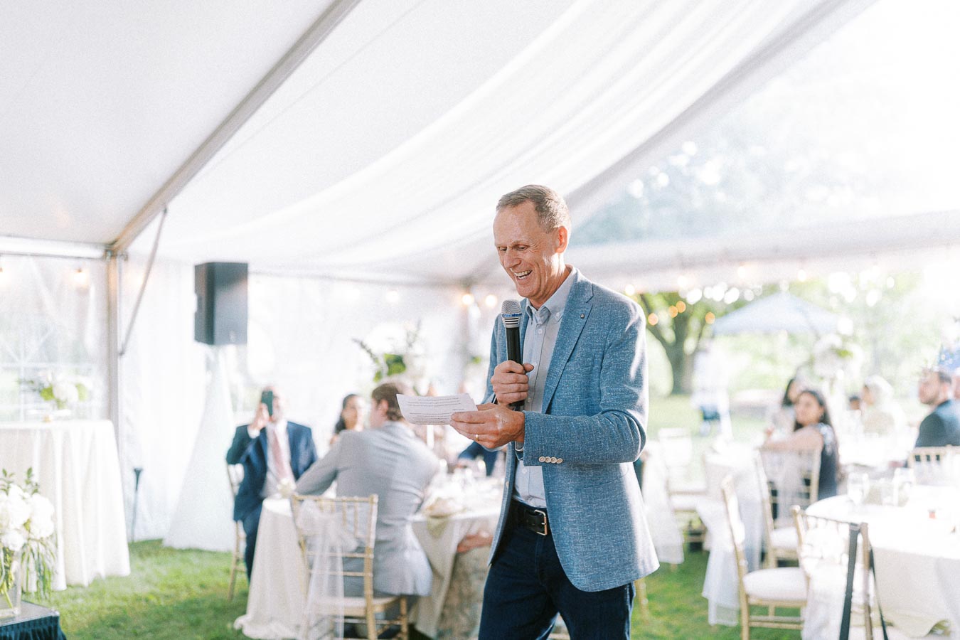 A man in a blue blazer holding a microphone gives a speech in a bright, outdoor event tent with guests seated at elegantly decorated tables.