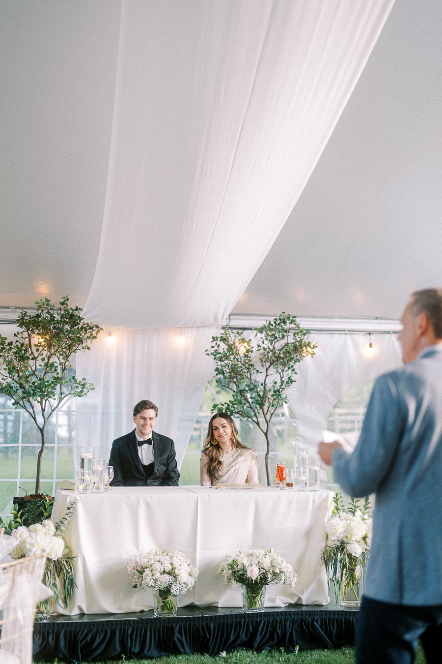 Wedding couple sitting at a decorated table under a tent, smiling during a speech. The elegant setting includes lush greenery, white floral arrangements, and soft lighting, creating a romantic atmosphere.
