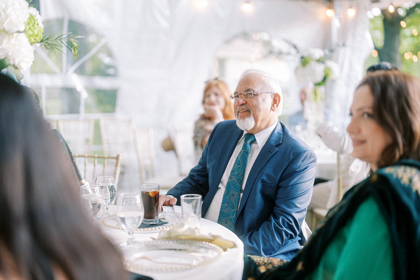 Elderly man in a blue suit and patterned tie sitting at an elegantly set table during an outdoor event, surrounded by guests in a decorated tent setting.