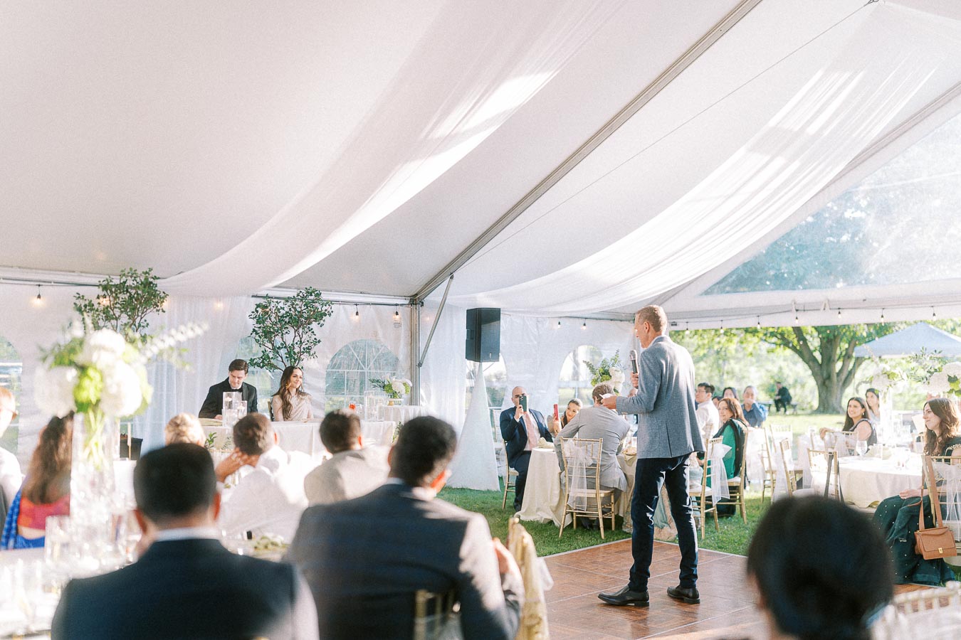 Outdoor wedding reception under a white tent with guests seated at tables and a person giving a speech with a microphone.