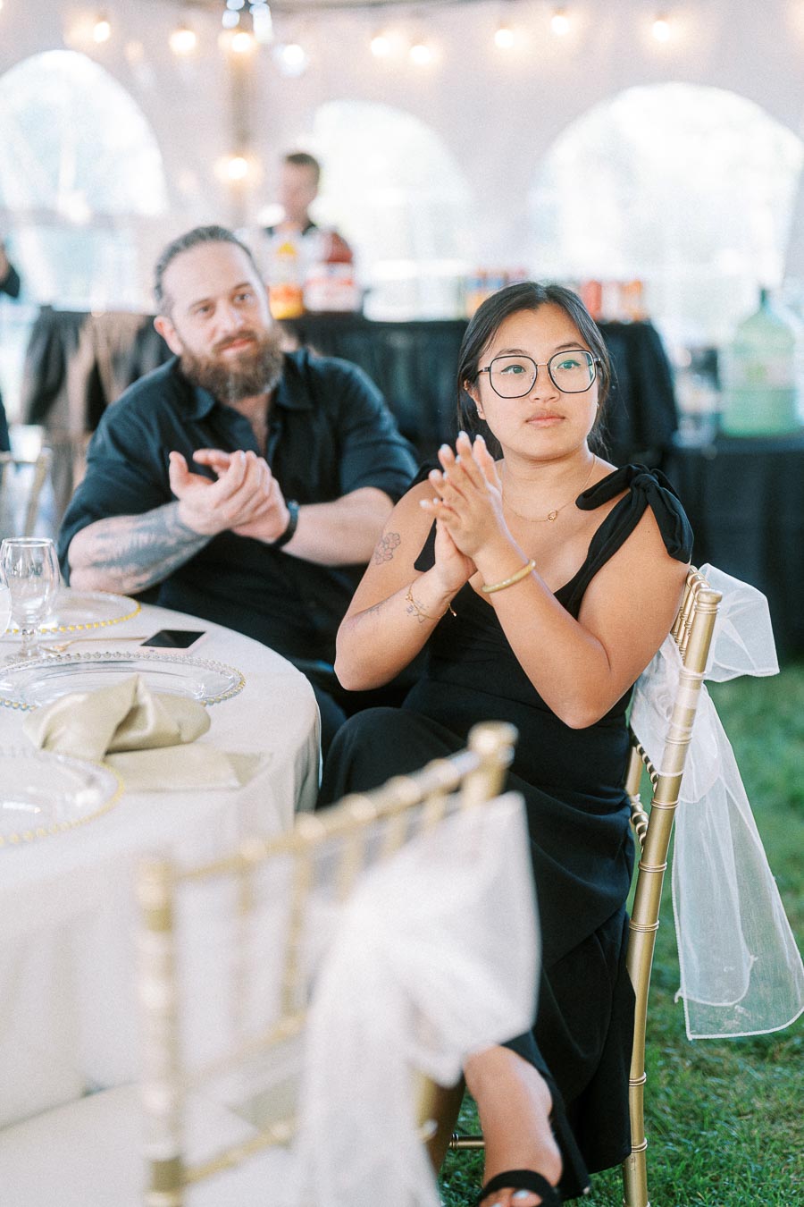 A woman and a man sitting and clapping at an elegantly decorated table during an outdoor event or wedding reception.