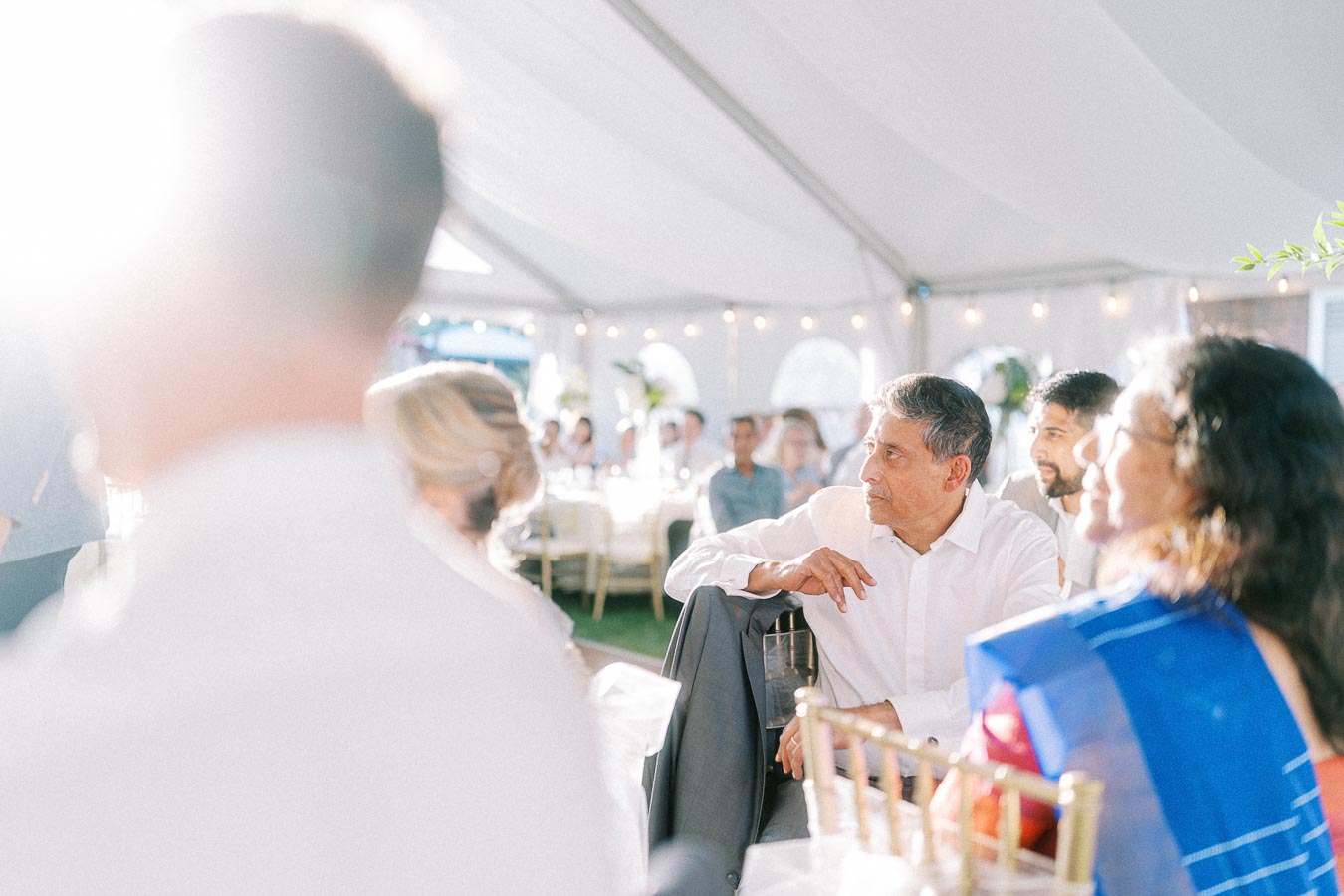 A diverse group of people enjoying a sunlit social gathering under a decorated tent, featuring elegant attire and vibrant setting elements.