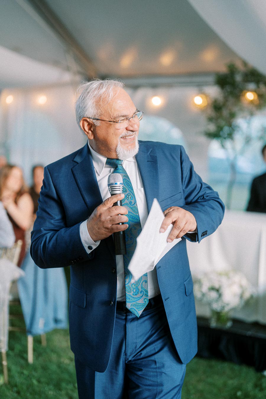 Elderly man in a blue suit speaking into a microphone at an outdoor event, smiling and holding a piece of paper.