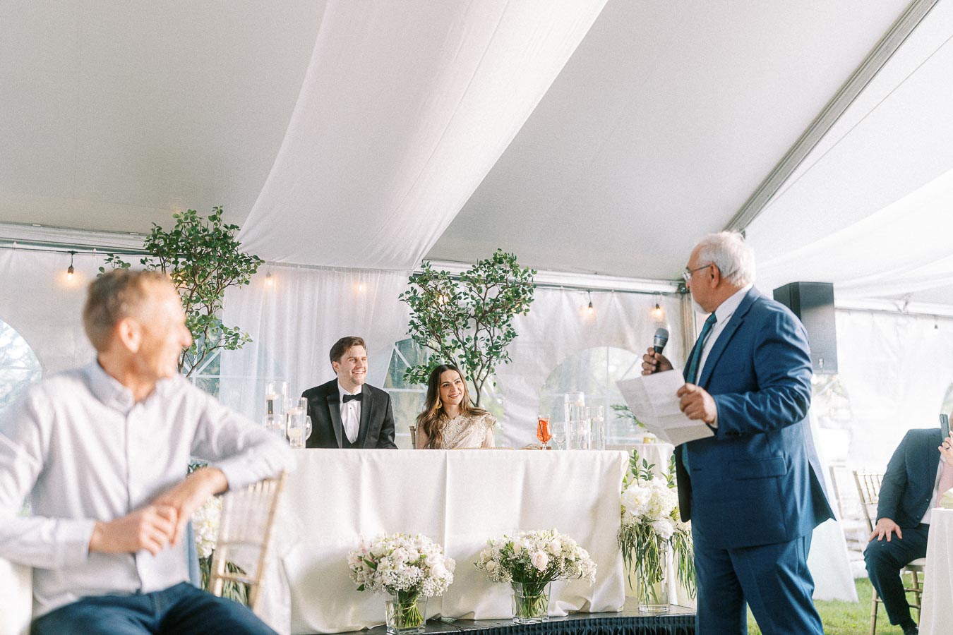 Wedding reception with a toast from an elderly man in a blue suit, addressing a smiling couple seated at a decorated head table under a tent, surrounded by floral arrangements and greenery.