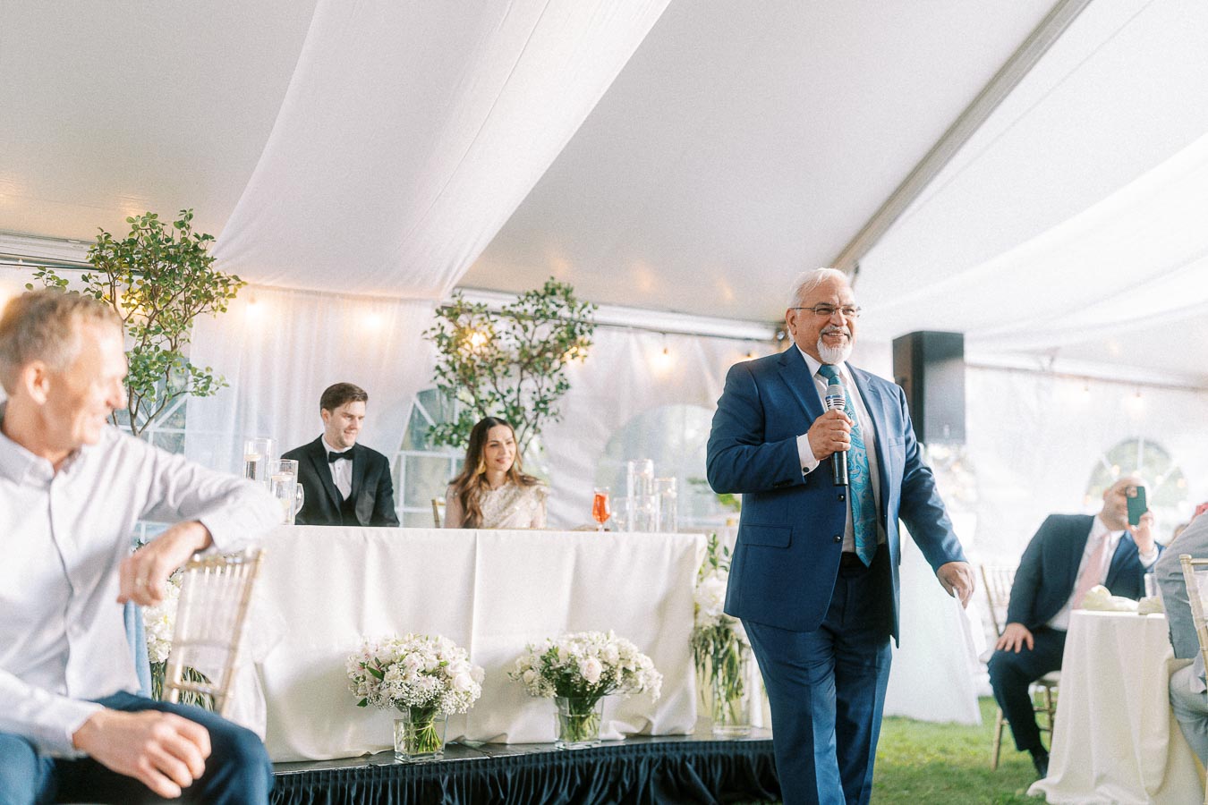 Elderly man in a blue suit giving a speech at a wedding reception inside a decorated tent, with the bride and groom seated at a table adorned with floral arrangements.