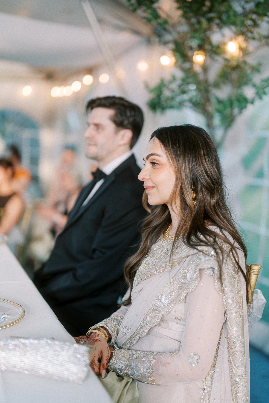 Elegant wedding guests sitting at a reception, with a woman in an embellished saree and a man in a tuxedo, under ambient string lights.