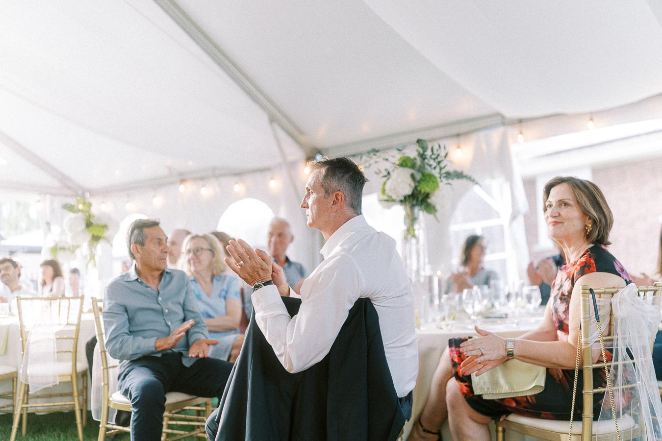 Guests seated under a white canopy at an elegant event, engaging in conversation and enjoying the ambiance. The festive setting features decorative lighting and floral centerpieces on the tables.