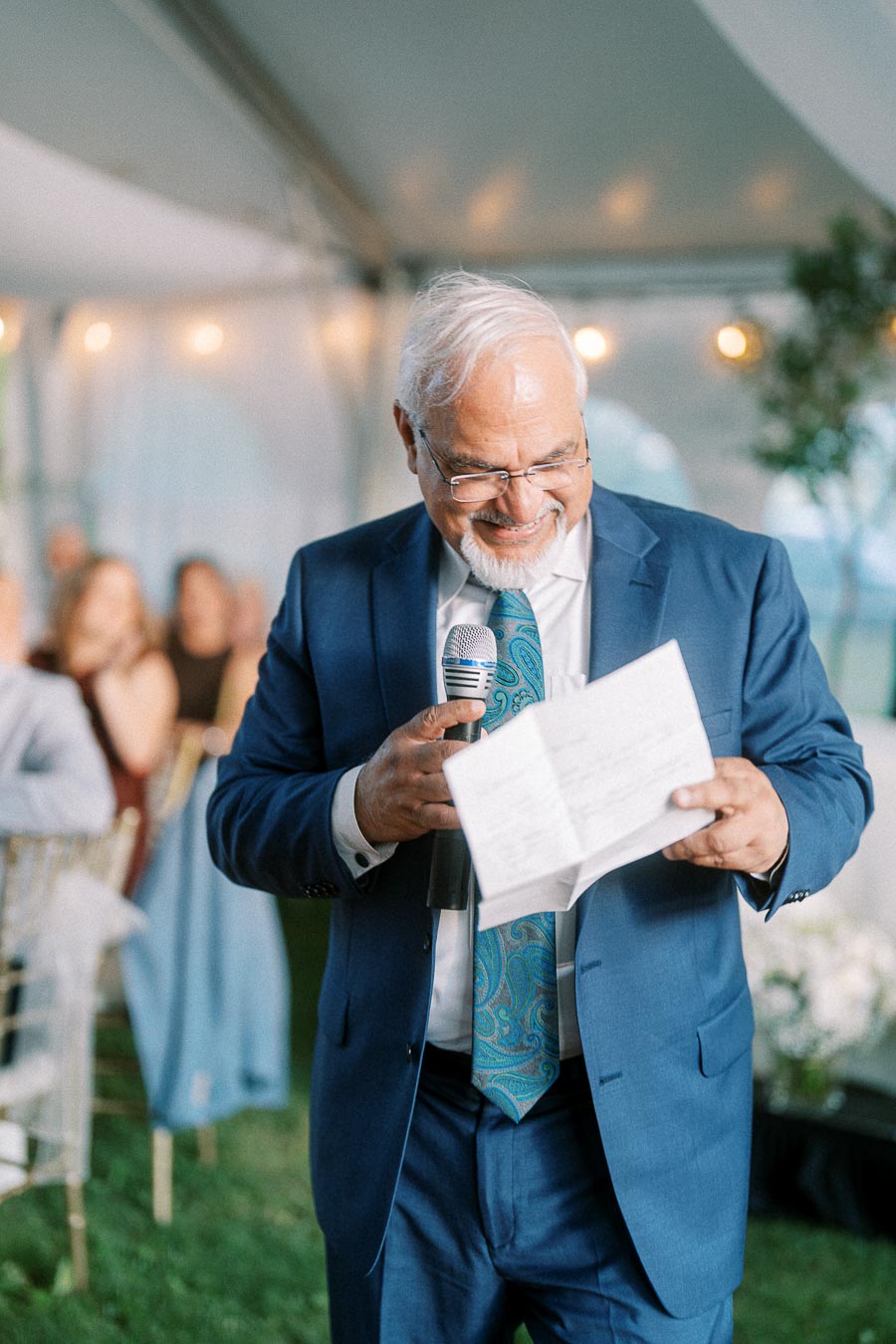 Elderly man in a blue suit giving a speech at an event while smiling, holding a microphone and reading from a paper.