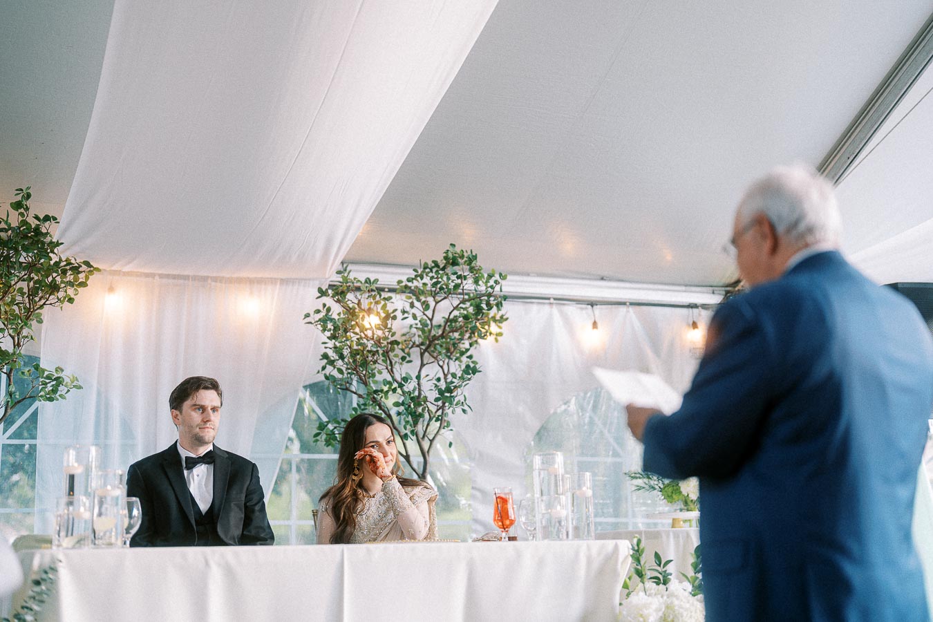 Wedding reception speech with bride and groom sitting at the head table under a decorated tent, featuring elegant greenery and thoughtful lighting.