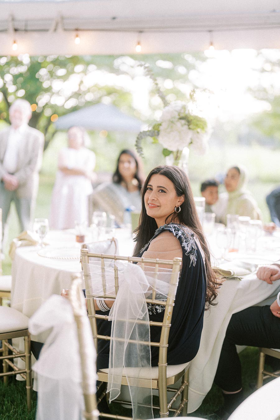 Elegant outdoor wedding reception with women in traditional attire seated at a beautifully decorated table, surrounded by lush greenery and soft, ambient lighting.