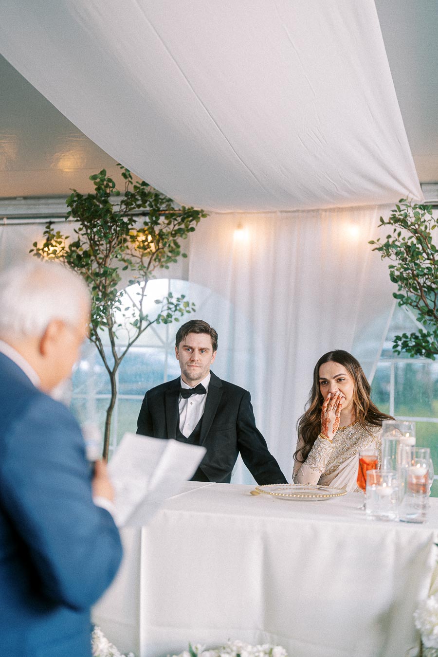 Wedding ceremony with a couple dressed elegantly, man in a black suit and woman in a traditional outfit with henna on her hands, listening to a speech under a white canopy adorned with greenery.