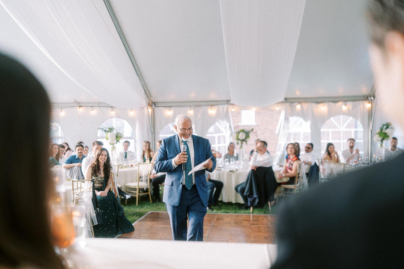 Elderly man in a blue suit giving a speech at a wedding reception under a white tent, surrounded by attentive guests seated at round tables, with string lights and floral decorations enhancing the elegant ambiance.