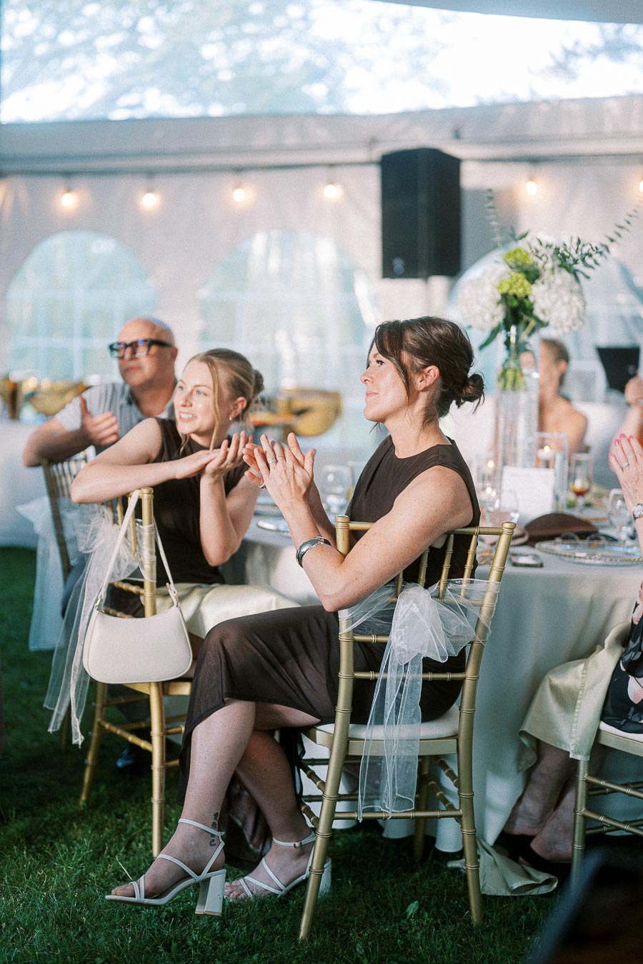 Guests seated and applauding at an elegant outdoor wedding reception, with women in formal dresses and decorative chairs covered in white fabric, set under a canopy with ambient lighting.