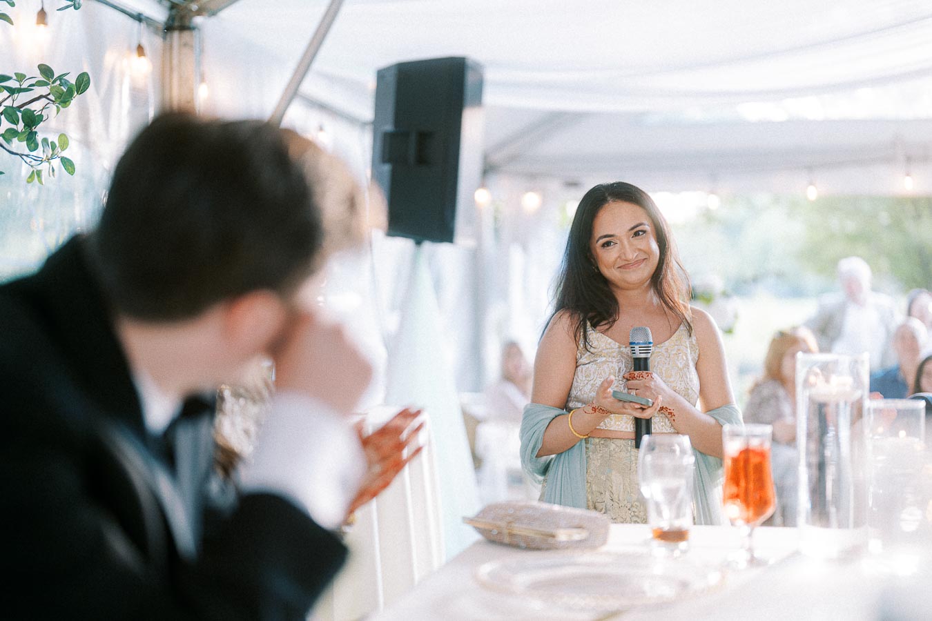 Elegant woman giving a heartfelt speech at a wedding reception, holding a microphone and smiling warmly, with a blurred background of guests and decorative lights in a marquee setting.