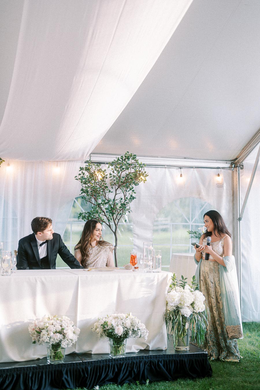 Wedding reception in a decorated tent with a couple seated at a table, a guest speaking with a microphone, elegant floral arrangements, and soft ambient lighting.