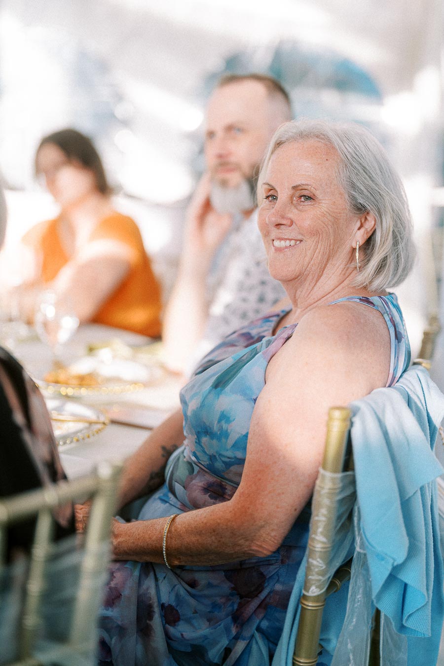 Elegant older woman smiling while seated at a wedding reception, wearing a floral dress and surrounded by other guests in a softly lit atmosphere.