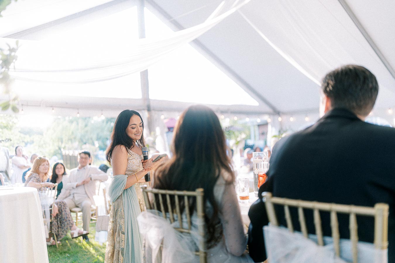 A woman delivers a heartfelt speech at an outdoor wedding reception, surrounded by attentive guests seated under a decorated tent with natural lighting.