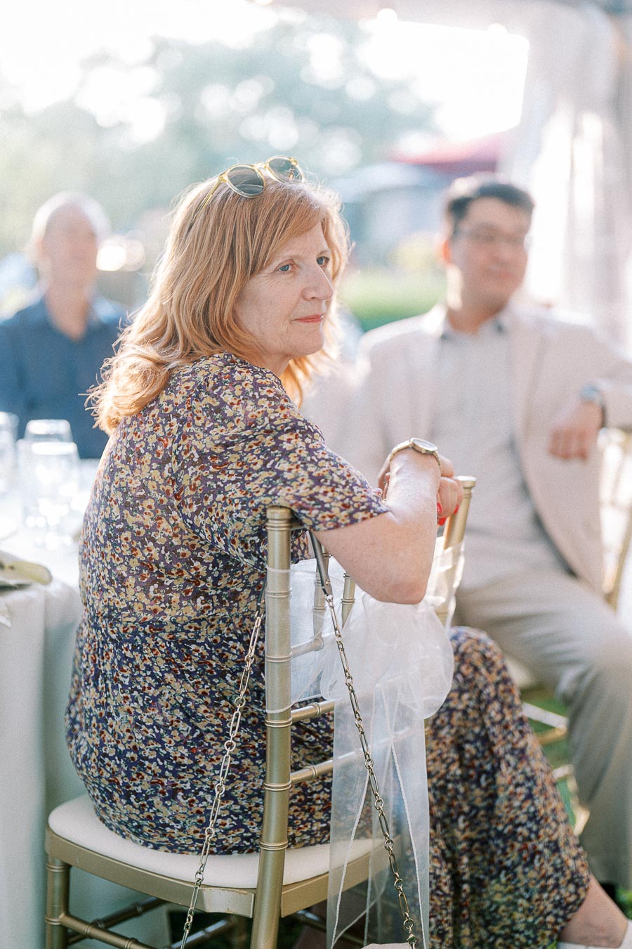 A woman seated at an outdoor event wearing a floral dress, with sunglasses resting on her head, attentively looking ahead, surrounded by blurred guests and bright daytime lighting.