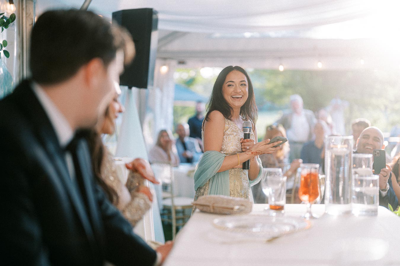 Smiling woman giving a speech at an outdoor event, holding a microphone and phone, with guests seated around in a sunlit setting.