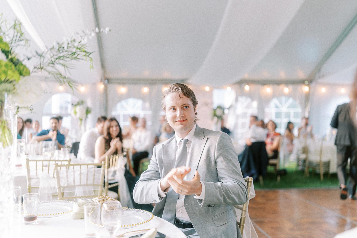 A man in a light gray suit clapping at a wedding reception inside a decorated tent, surrounded by elegantly dressed guests.