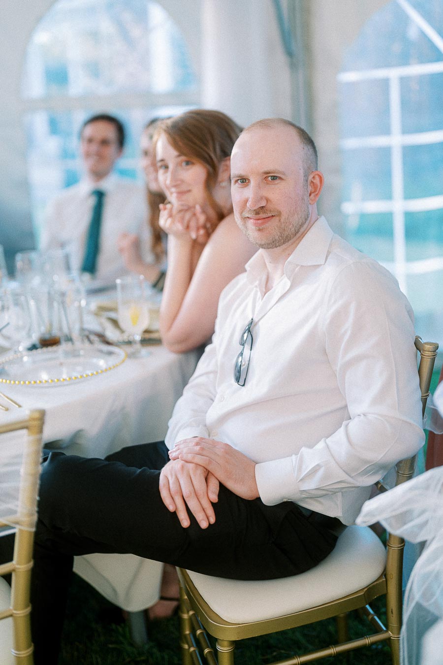 A man in a white shirt sitting at an elegantly decorated table during a wedding reception, with other guests visible in the background.
