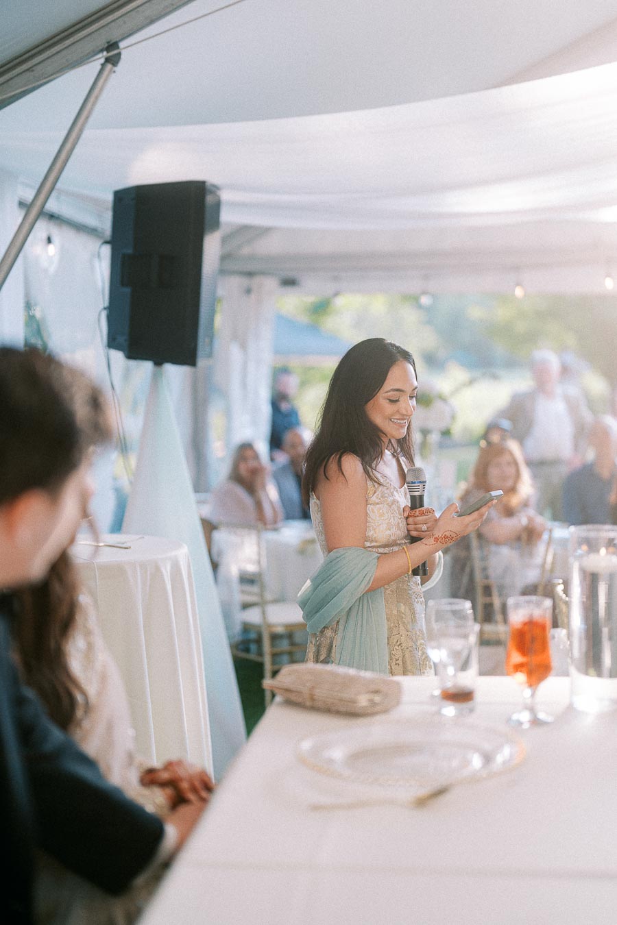Woman giving a speech at an outdoor wedding reception, wearing a light-colored traditional dress with a blue shawl, holding a microphone while reading from her phone. The setting is elegantly decorated with guests seated at tables.