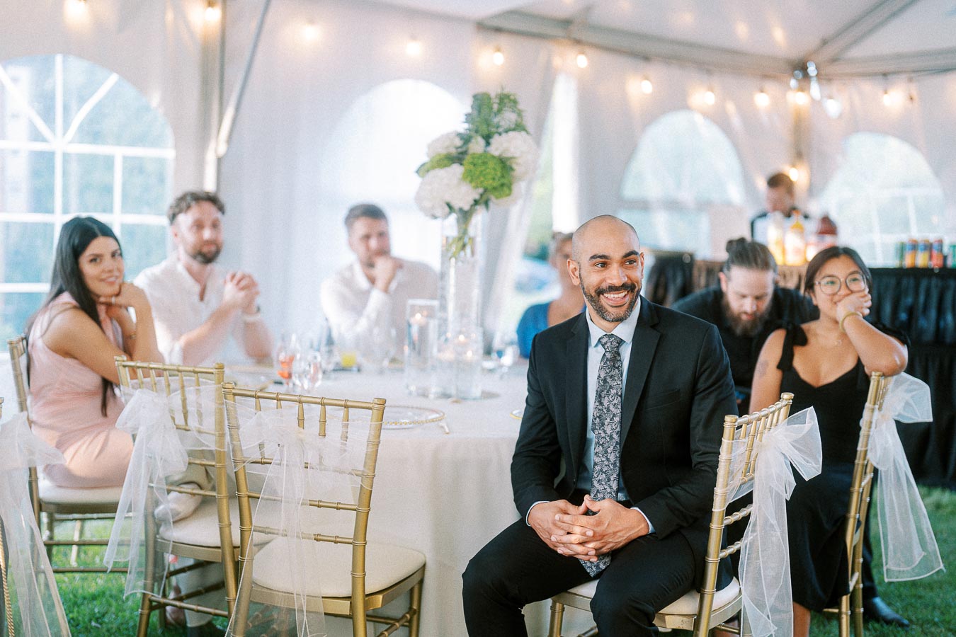 Group of people seated at a decorated table inside a tent during a formal event, with elegant chairs and ambient lighting.