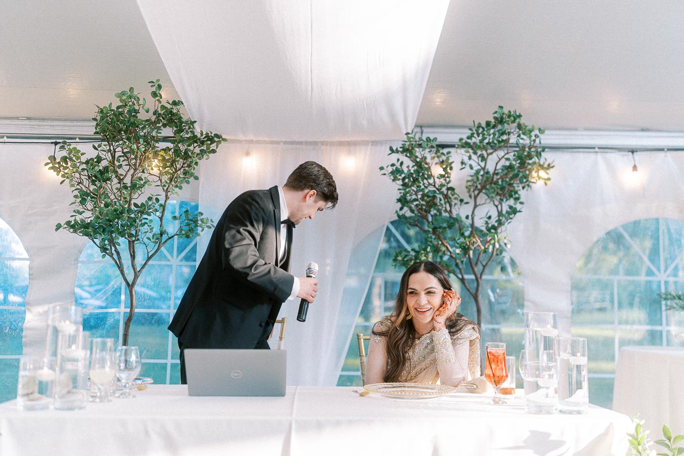 Man in a suit holding a microphone next to a smiling woman in an elegant dress seated at a decorated table with a laptop, set within a tent adorned with greenery and warm lighting.