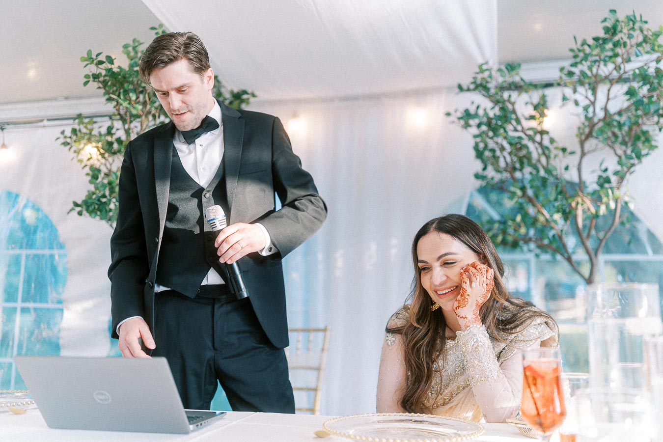 A man in a tuxedo holds a microphone while standing next to a woman in a decorative outfit with henna on her hand, smiling as they look at a laptop during a wedding event.