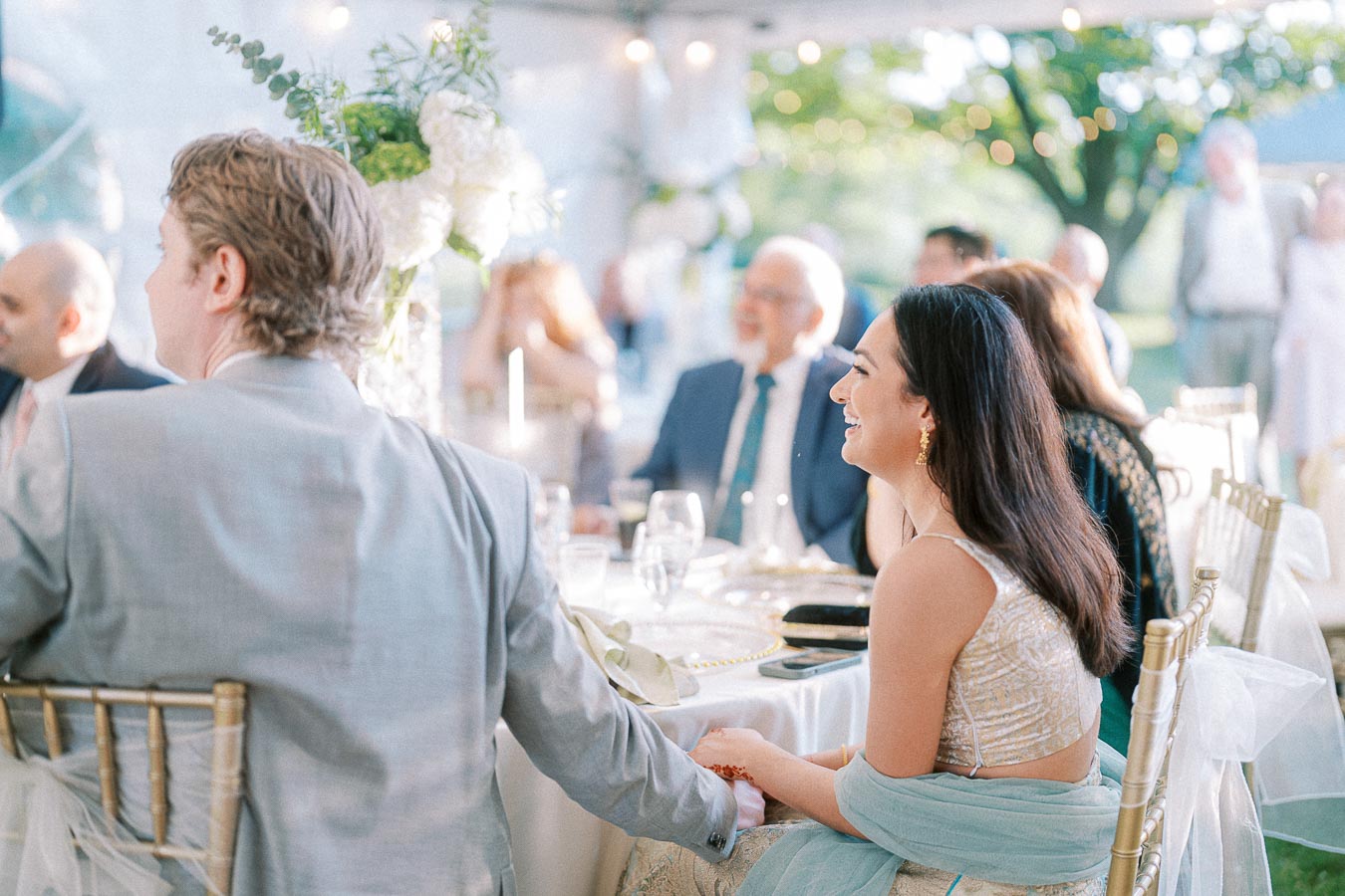 A joyful outdoor wedding reception with elegantly dressed guests sitting around a decorated table. The seating area is adorned with lush floral arrangements and soft, ambient lighting under a canopy.