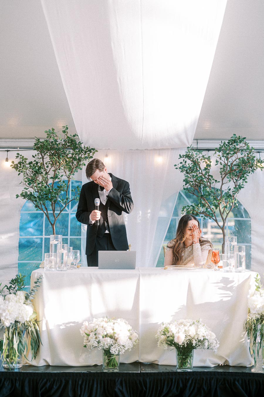 A groom delivering an emotional speech during a wedding reception inside a beautifully decorated tent, with the bride visibly touched while seated beside him at the table adorned with elegant floral arrangements.