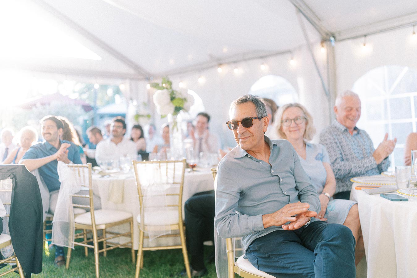 A group of guests seated at an elegantly decorated wedding reception, clapping and enjoying the event under a sunlit tent with white drapery and soft lighting.