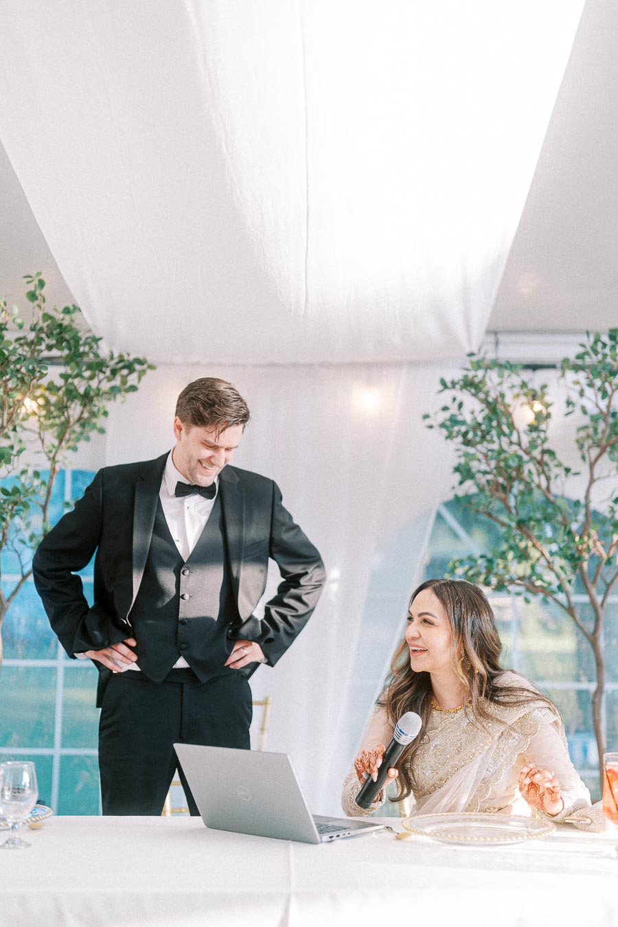 A woman in a traditional outfit holding a microphone sits beside a laptop on a table, smiling up at a man in a tuxedo standing next to her, during a joyful indoor celebration with natural light and greenery in the background.
