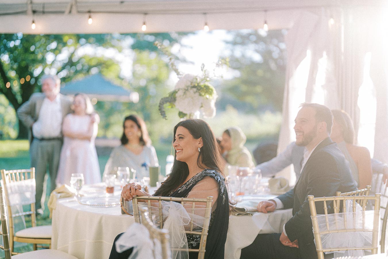 Elegant outdoor wedding reception with guests smiling at a beautifully decorated table under a tent, featuring string lights, floral centerpieces, and a sunlit garden setting.
