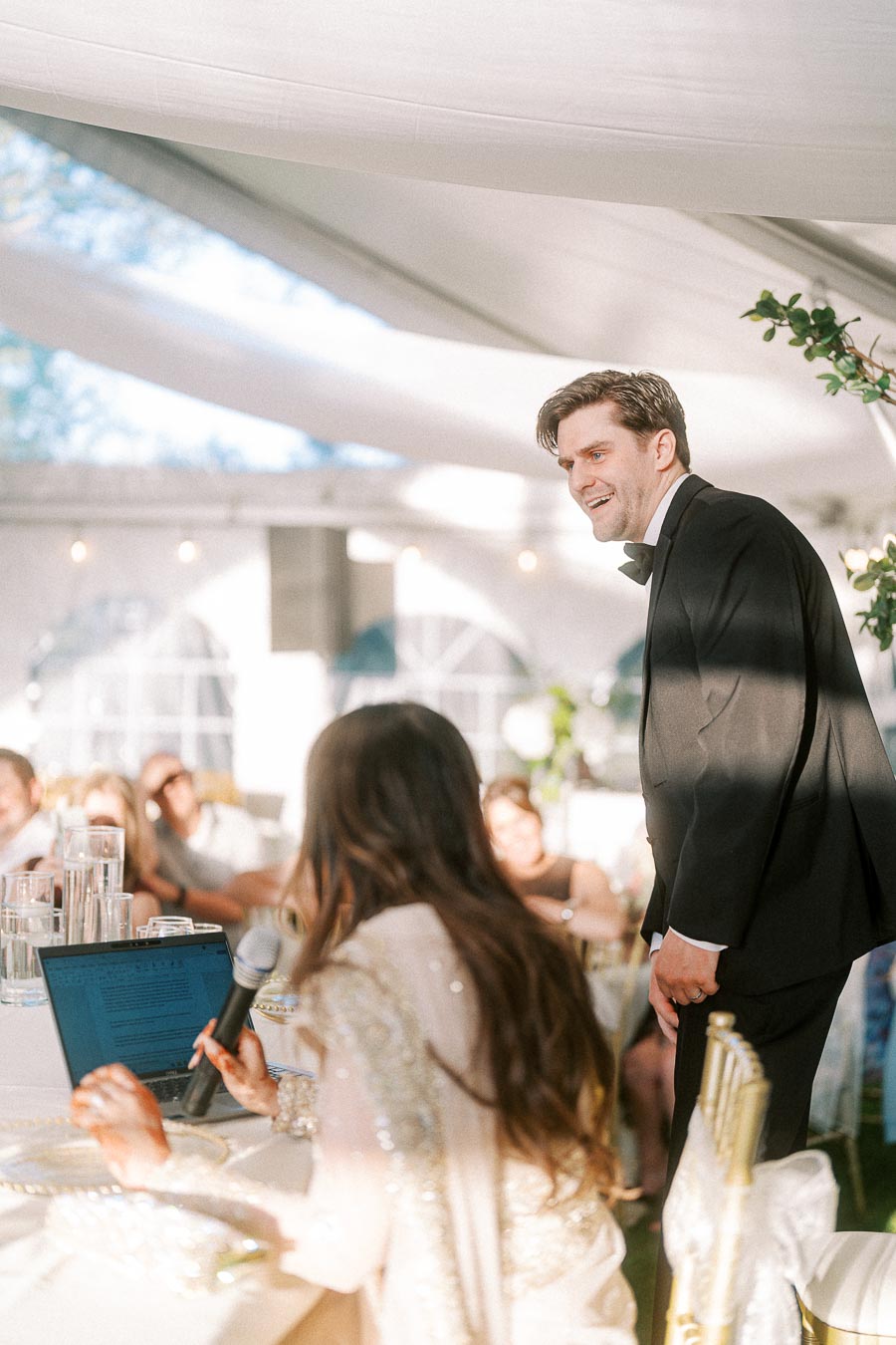 A groom in a tuxedo smiling during a wedding reception speech, under a beautifully lit tent with guests seated at tables in the background.
