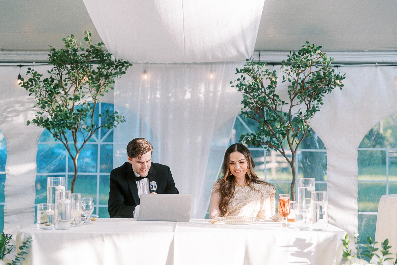 Bride and groom seated at a beautifully decorated wedding reception table, under a white tent adorned with greenery, with the groom speaking into a microphone and the bride smiling.