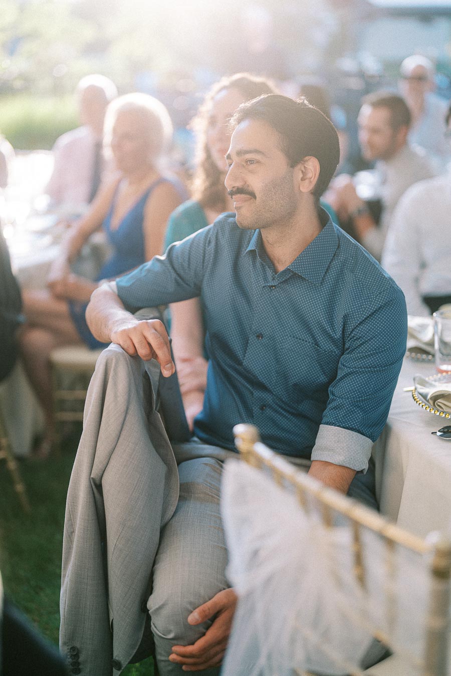 Man in blue shirt sitting attentively at a sunny outdoor event, holding a light gray suit jacket, surrounded by other seated guests.